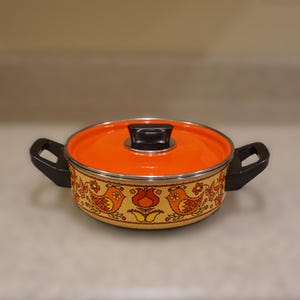 Puede incluir: Una olla de cocina vintage naranja y amarilla con tapa y asas negras. La olla presenta una banda decorativa con diseños de pájaros y flores. La tapa tiene un asa negra.