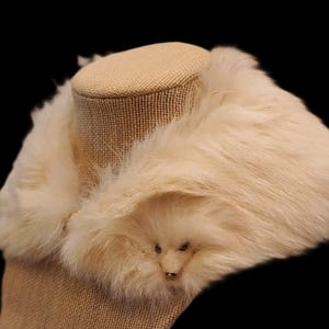May include: Cream-colored faux fur collar with a small, decorative animal face detail. The collar is displayed on a mannequin head with a burlap-like texture, set against a black background. A cozy accessory.