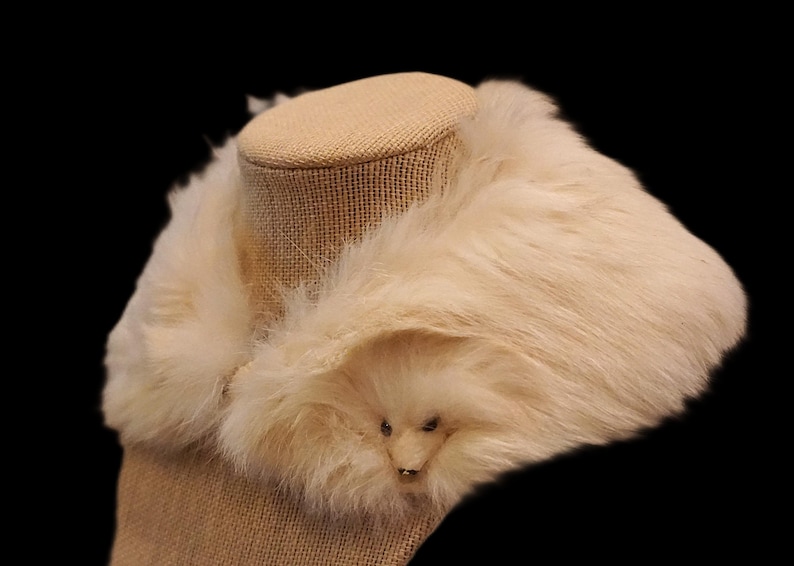May include: Cream-colored faux fur collar with a small, decorative animal face detail. The collar is displayed on a mannequin head with a burlap-like texture, set against a black background. A cozy accessory.