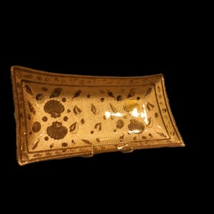 Puede incluir: Una bandeja de servir rectangular de vidrio de color dorado con un patrón decorativo de conchas marinas y hojas. La bandeja es rectangular y tiene un borde ligeramente curvado.