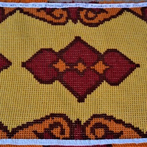 May include: A yellow and red needlepoint canvas with a geometric pattern. The canvas is framed with white and orange fringe.