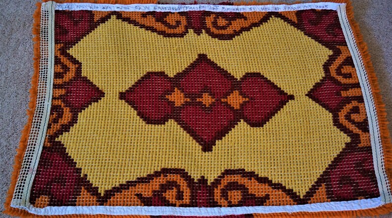 May include: A yellow and red needlepoint canvas with a geometric pattern. The canvas is framed with white and orange fringe.
