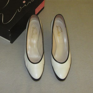 May include: Pair of white leather pumps with black trim. The shoes have a pointed toe and a low heel. The brand name "Evviva" is written on the inside of each shoe.