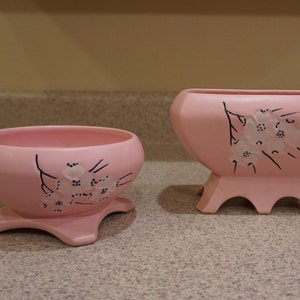 May include: Two pink ceramic planters with black floral designs. The larger planter has a rectangular shape with four legs. The smaller planter has a round shape with three legs.