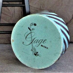 May include: A blue and black striped hat box with the words "Jage Hats" printed on the top. The box is sitting on a brown surface.