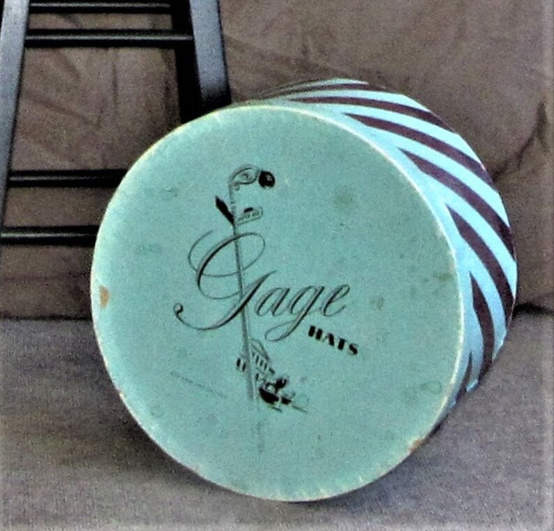 May include: A blue and black striped hat box with the words "Jage Hats" printed on the top. The box is sitting on a brown surface.