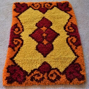 May include: A small, rectangular rug with a geometric pattern in shades of orange, yellow, and red. The rug has a fringed edge and is made of a soft, plush material.