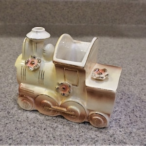 May include: A white ceramic train engine planter with gold accents and pink flowers. The planter has a rectangular opening for planting.