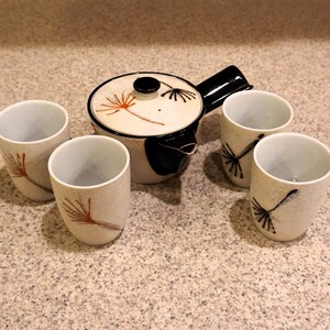 May include: A white teapot with a black handle and a black lid, with a dandelion design. There are four white teacups with a dandelion design, all on a beige surface.