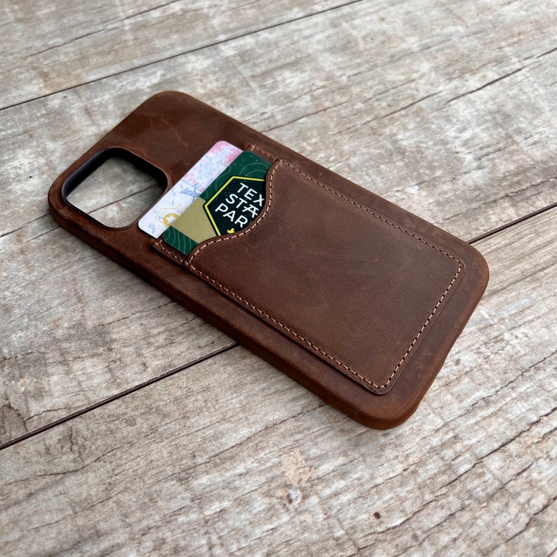 Op de afbeelding: Een bruine leren telefoonhoes met een kaartslot. Het kaartslot heeft een gestikte rand en een kaart is zichtbaar in het slot. De kaart heeft de tekst "Texas State Park" erop.