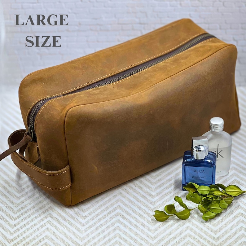 May include: A large brown leather toiletry bag with a zipper closure and a handle. The bag is sitting on a white and gray patterned surface.