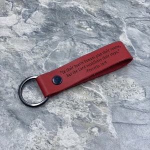 Peut inclure: Porte-cl&eacute;s en cuir rouge avec un anneau argent&eacute; et la citation "Dans leur c&oelig;ur, les humains planifient leur route, mais le Seigneur &eacute;tablit leurs pas." -Proverbes 16:9 grav&eacute;e dessus.