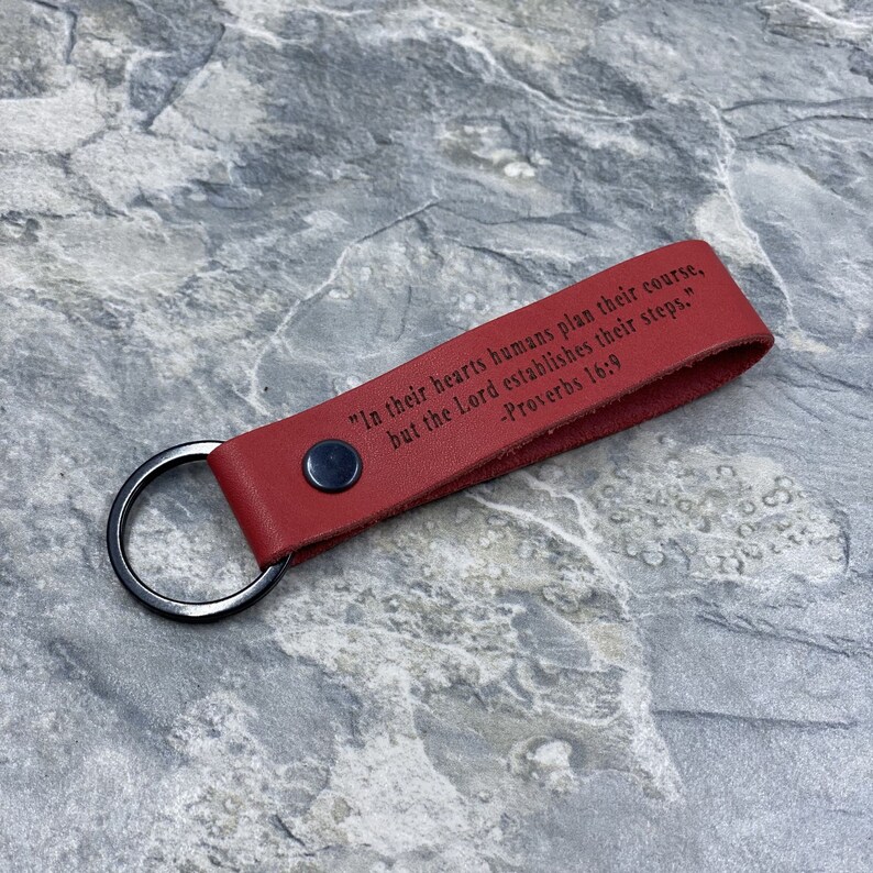 Peut inclure: Porte-cl&eacute;s en cuir rouge avec un anneau argent&eacute; et la citation "Dans leur c&oelig;ur, les humains planifient leur route, mais le Seigneur &eacute;tablit leurs pas." -Proverbes 16:9 grav&eacute;e dessus.