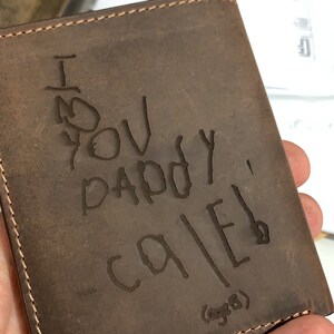May include: A brown leather wallet with a stitched edge. The wallet has a message written on it in black ink that says "I love you Daddy - Caleb (age 8)"