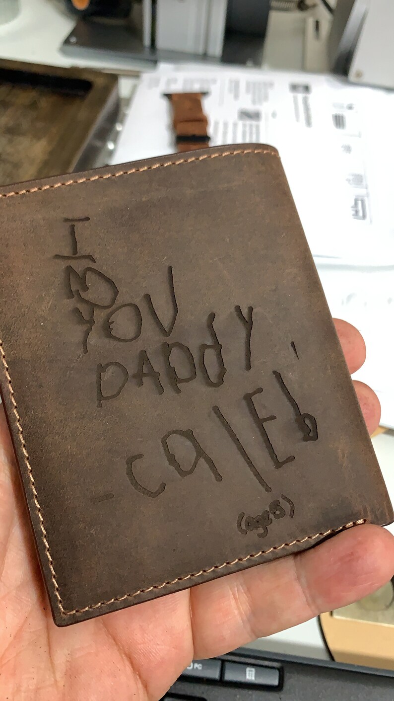 May include: A brown leather wallet with a stitched edge. The wallet has a message written on it in black ink that says "I love you Daddy - Caleb (age 8)"