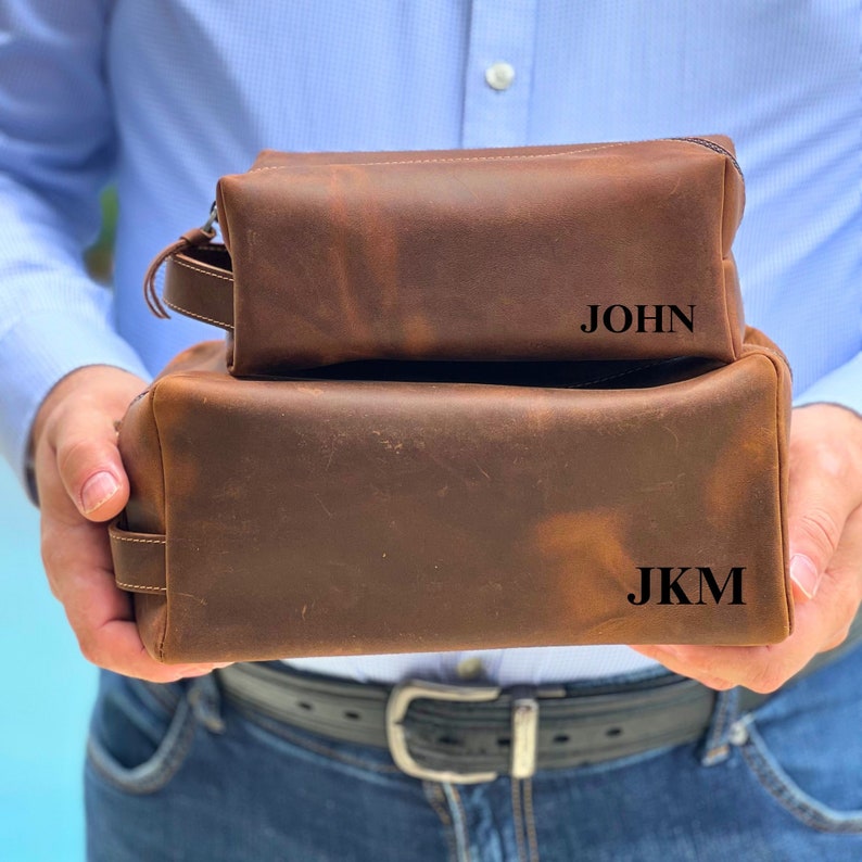 May include: Two brown leather toiletry bags with zipper closures. The smaller bag has the name "JOHN" embossed on the front. The larger bag has the initials "JKM" embossed on the front.