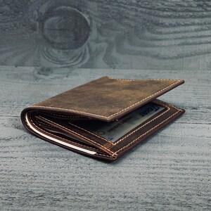 May include: A brown leather wallet with a stitched edge and a visible card slot. The wallet is folded and lying on a gray wooden surface.