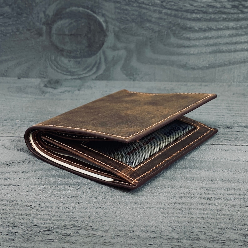May include: A brown leather wallet with a stitched edge and a visible card slot. The wallet is folded and lying on a gray wooden surface.