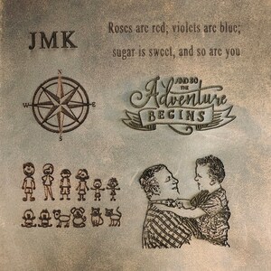 May include: A brown leather journal with the initials "JMK" embossed on the front cover. The journal also features a compass, a family of stick figures, and a father and son embracing. The text "And so the adventure begins" is embossed on the cover. The journal has a quote about roses, violets, sugar, and sweetness.