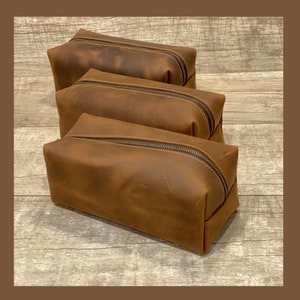 May include: Three brown leather zippered pouches stacked on top of each other. The pouches are made of a distressed leather with a rustic look.