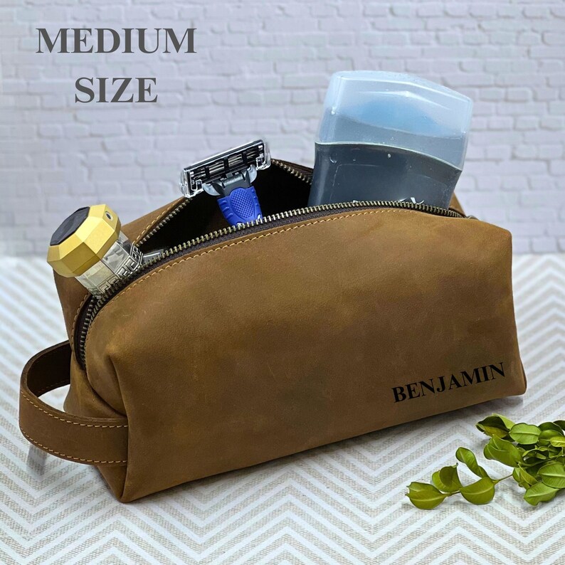 May include: A brown leather toiletry bag with a zipper closure. The bag is personalized with the name "BENJAMIN". The bag is open and contains a razor, a deodorant stick, and a small bottle.