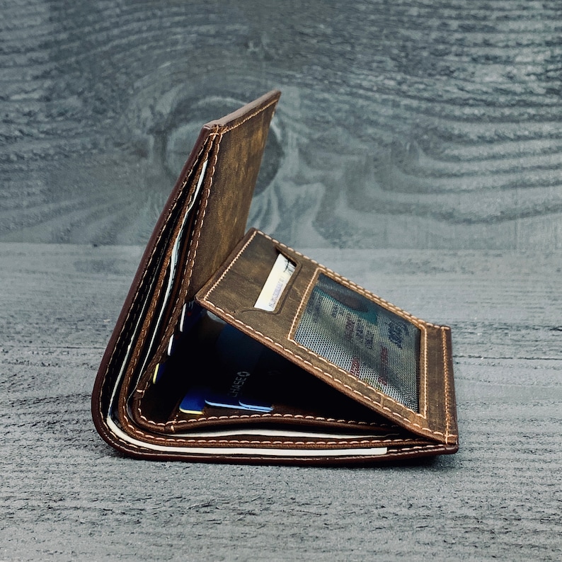 May include: A brown leather wallet with multiple card slots and a clear ID window. The wallet is folded closed and is lying on a gray wooden surface.
