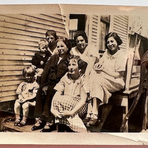 May include: A black and white photo of a family of seven posing outside a house. The family is standing and sitting on the porch steps. There is a hand pump in the background.