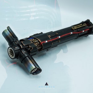 Puede incluir: Un sable de luz desmontado con un acabado negro y cobre. El sable de luz está formado por varias piezas, incluyendo una empuñadura, una hoja y una célula de energía. El sable de luz está en estado desmontado, con las piezas sobre una superficie blanca.