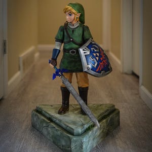 May include: An action figure of Link from the Legend of Zelda video game series. He is wearing a green tunic and holding a silver sword and a blue and red shield. He is standing on a green and brown stone base.