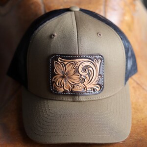 May include: A green and black trucker hat with a brown leather patch featuring a tooled floral design.