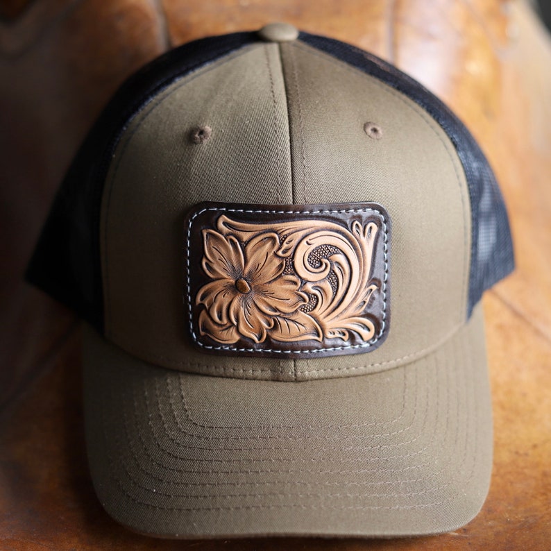 May include: A green and black trucker hat with a brown leather patch featuring a tooled floral design.
