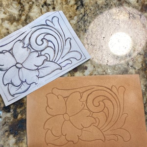May include: A close-up of a leather craft project. A floral design is carved into a piece of brown leather. A paper pattern with the same design is visible in the background.