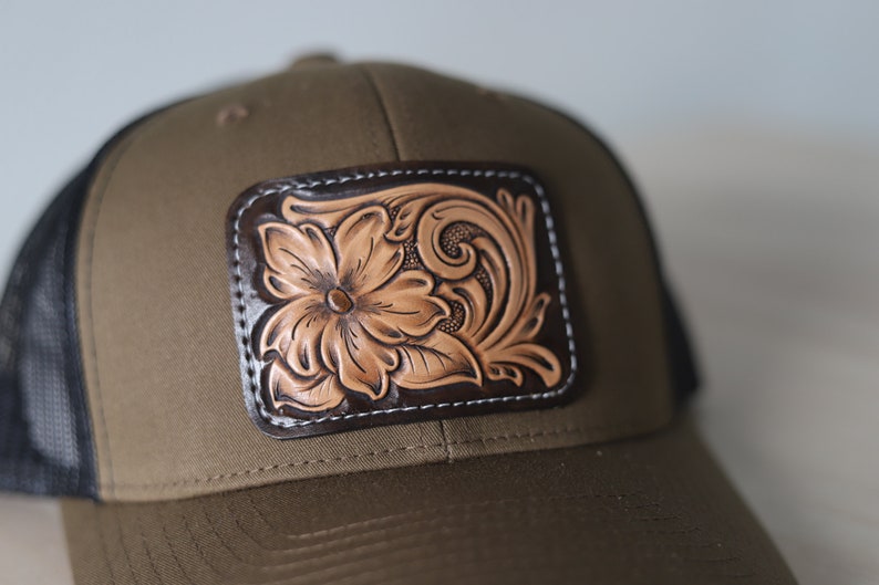 May include: A brown and black trucker hat with a leather patch featuring a tooled floral design.
