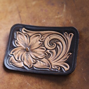 May include: A brown leather patch with a tooled design of a flower and scrollwork. The flower is in a light brown color and the scrollwork is in a darker brown color.