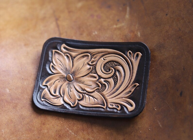 May include: A brown leather patch with a tooled design of a flower and scrollwork. The flower is in a light brown color and the scrollwork is in a darker brown color.