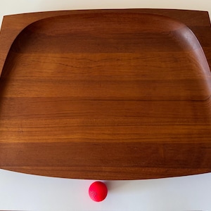 May include: A rectangular wooden tray with a curved edge. The tray is made of a warm brown wood with visible grain. A small red ball is at the bottom of the tray.