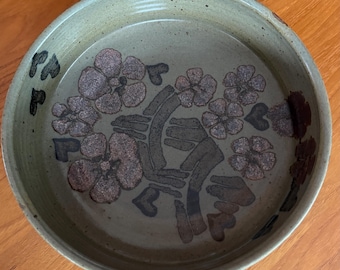 May include: A round, ceramic serving dish with a floral design. The dish has a light green base with brown and black floral and leaf patterns. The rim is slightly raised, and the dish sits on a wooden surface.