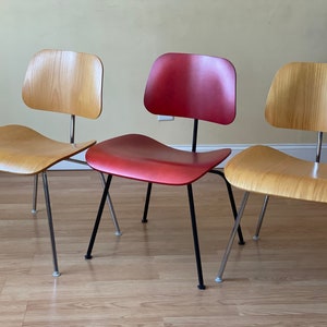 Peut inclure: Trois chaises avec des sièges et des dossiers en bois. Une chaise a un siège rouge et des pieds noirs, tandis que les deux autres ont des sièges en bois naturel et des pieds chromés.