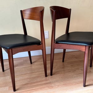 Peut inclure: Deux chaises modernes du milieu du siècle avec des cadres en bois foncé et des assises en cuir noir. Les chaises ont des dossiers incurvés et des pieds effilés, mettant en valeur un design classique. Les chaises sont placées contre un mur neutre.