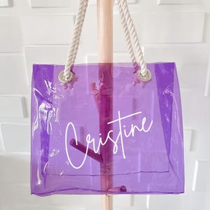 May include: A transparent purple tote bag featuring the name Cristine written in white cursive script. The bag has two thick white rope handles attached through silver metal grommets at the top, providing a clear view of the interior structure.