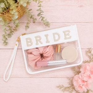 May include: A clear rectangular pouch with a white trim and a white wrist strap. The word BRIDE is spelled out in gold-toned, pearl-embellished letters across the top. Inside the pouch are a pink scrunchie, a small glass bottle, and a tube.