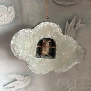 May include: A whimsical, handmade cloud-shaped ornament with a small, brown animal figure inside. The cloud is painted in shades of blue and white, with a textured appearance. It hangs from a natural twine string, suggesting a decorative or seasonal item.