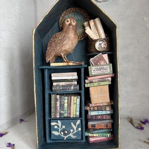 May include: A miniature diorama in the shape of a house, painted in teal with gold trim. It features a brown owl figurine, miniature books, and a small drawer with a floral design. A small globe and rolled scrolls are also visible.