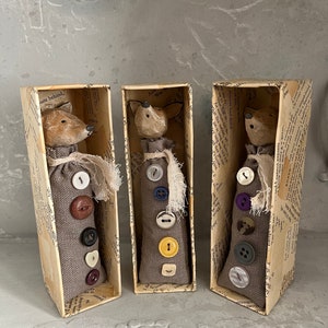 May include: Three whimsical animal figures in rectangular boxes. Each figure has a painted wooden head and a gray fabric body adorned with colorful buttons. The boxes are lined with aged paper, creating a vintage aesthetic. A decorative piece for a shelf.