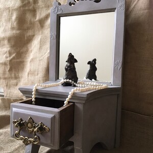 May include: A gray, miniature vanity with a mirror, drawer, and decorative carvings. A pearl necklace and two small statues are on the vanity. The drawer has a brass handle. The mirror reflects the statues.