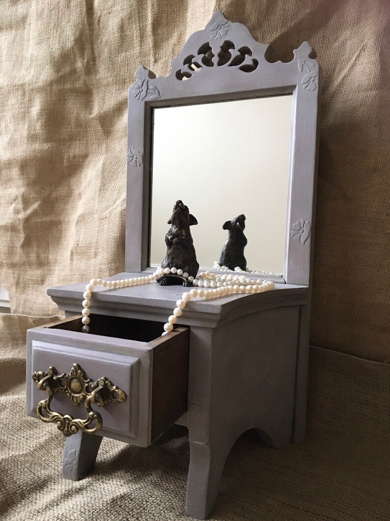May include: A gray, miniature vanity with a mirror, drawer, and decorative carvings. A pearl necklace and two small statues are on the vanity. The drawer has a brass handle. The mirror reflects the statues.