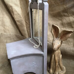 May include: A small, gray jewelry box with a mirror and a pearl necklace. The box has a decorative floral design and is next to a ceramic rabbit figurine. The background is a tan burlap.