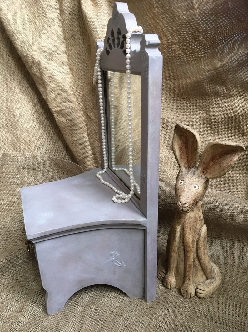 May include: A small, gray jewelry box with a mirror and a pearl necklace. The box has a decorative floral design and is next to a ceramic rabbit figurine. The background is a tan burlap.