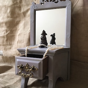 May include: A gray vanity mirror with a small drawer and decorative gold-coloured handle. The mirror has a carved top and is adorned with two small, dark-coloured figurines and a strand of pearls. The vanity is approximately 30 cm tall.