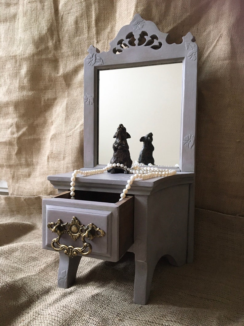 May include: A gray vanity mirror with a small drawer and decorative gold-colored handle. The mirror has a carved top and is adorned with two small, dark-colored figurines and a strand of pearls. The vanity is approximately 12 inches tall.